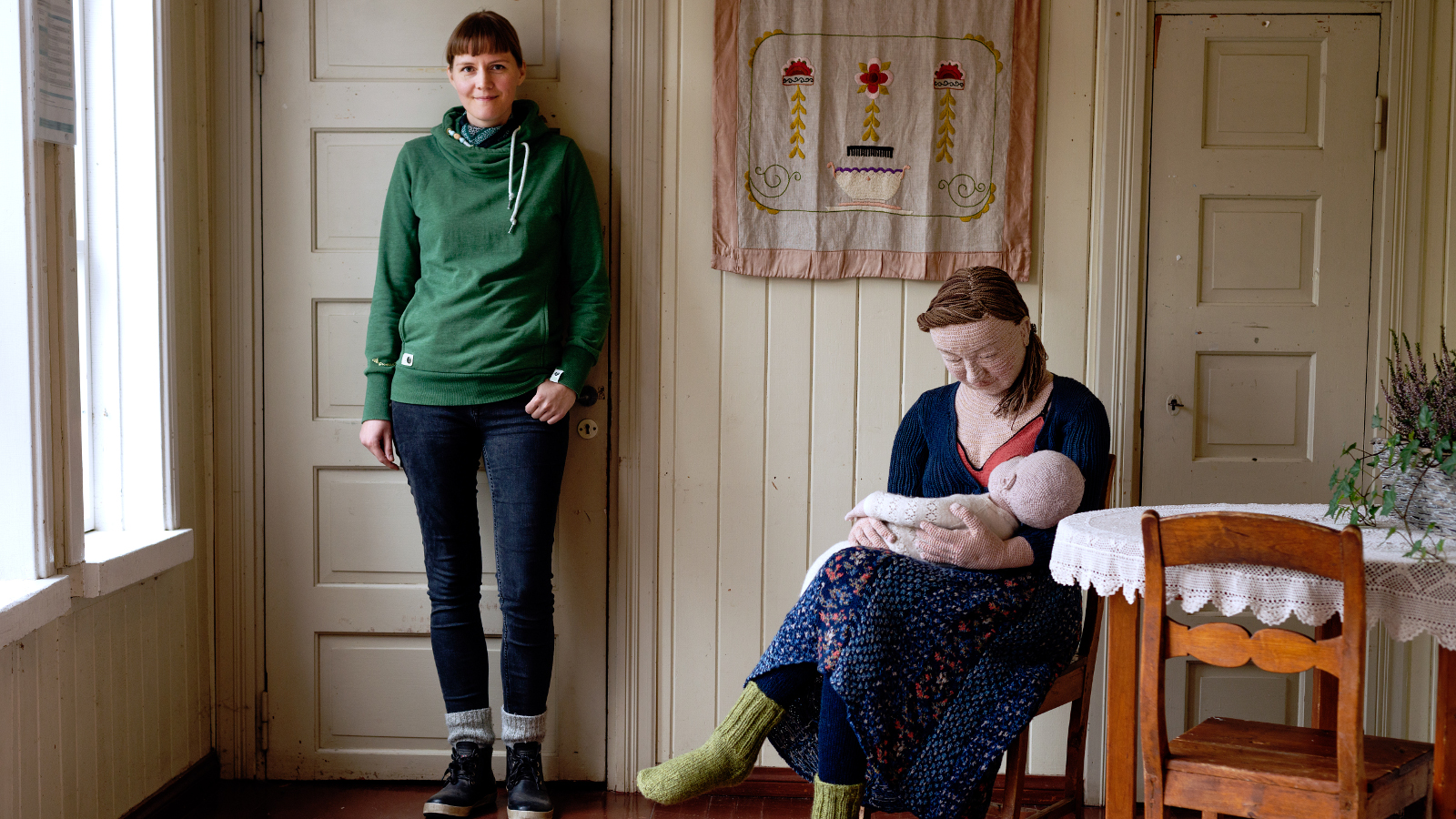 Kuvassa on Liisa Hietanen seisomassa huoneen seinään nojaten ja vierellä on tuolissa istumassa nainen, jolla on vauva sylissään. Nämä ovat virkattuja.
