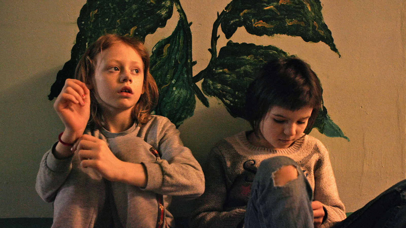 Kuvassa on kaksi tyttöä istumassa sohvalla. Kuvan sävy on ruskehtava. Takana on seinässä tumma kasvikuvio.