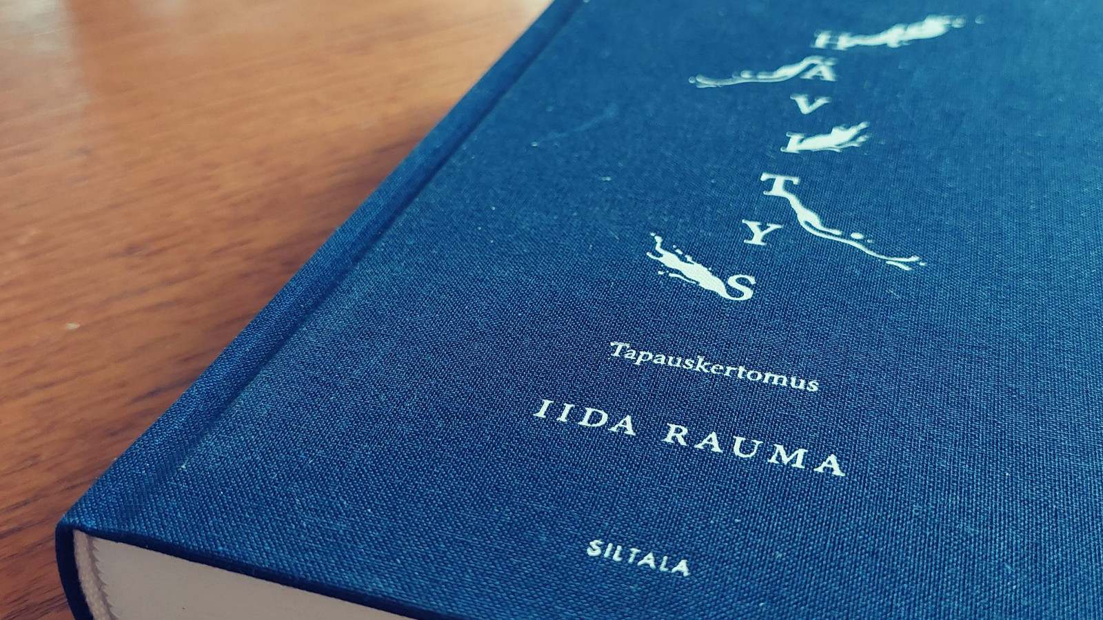 Kuvassa on sininen kirja vaalean ruskean pöydän kannella. Kirjan kannessa on sen nimi Hävitys -tapauskertomus valkoisella melko pienillä kirjaimilla, kirjoittaja Iida Rauma ja alhaalla kustantaja Siltala.