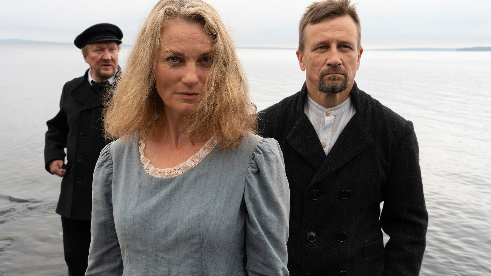 Kuvassa on etualalla harmaassa mekossa Jonna Järnefelt ja taustalla kaksi miestä mustissa takeissa.  He kaikki seisovat ulkona meren rannalla.