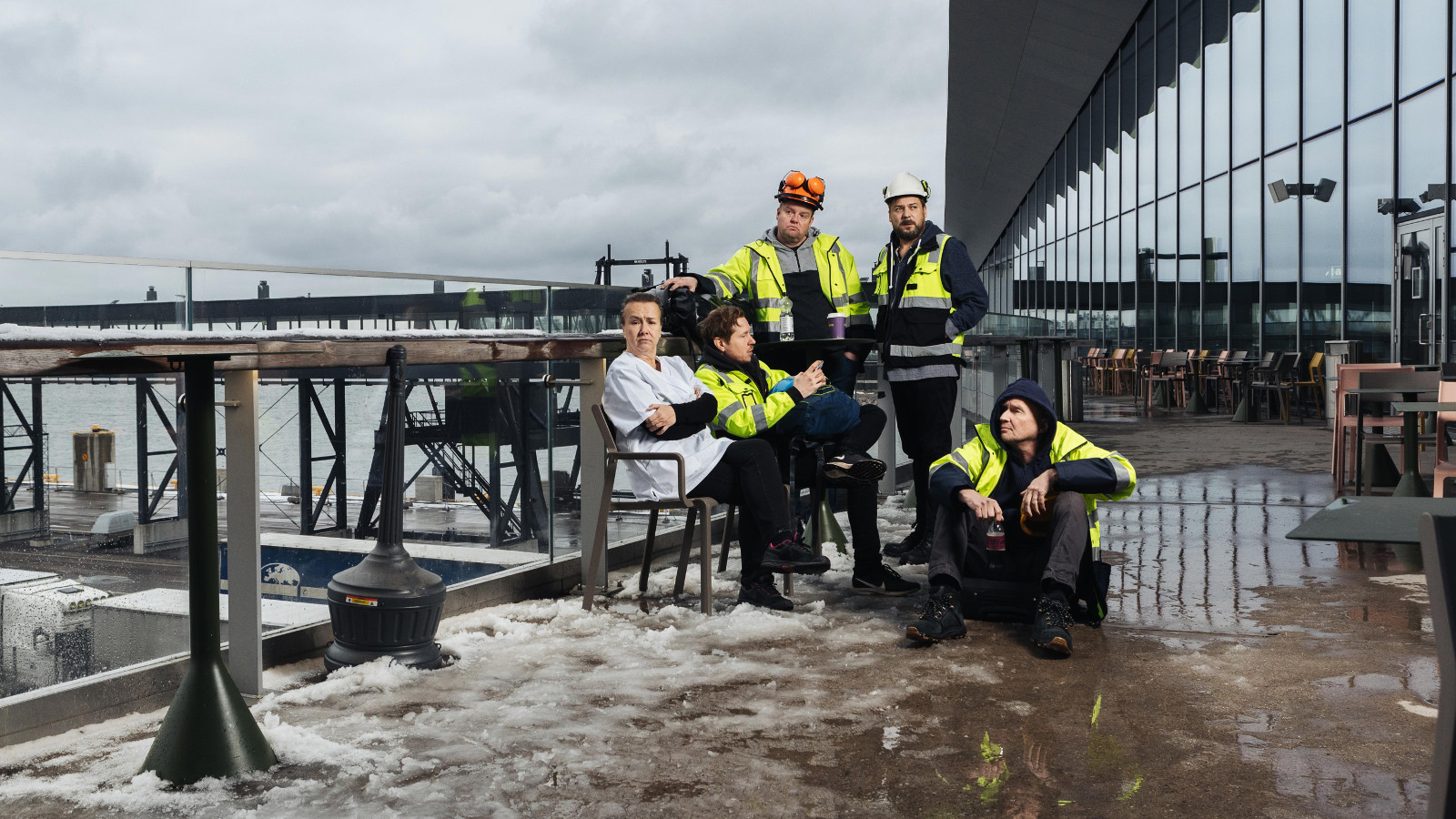 Kuvassa on vasemmalla rakennustyömaata ja vettä.  Oikealla on vinosti lasi-ikkunainen toimistorakennus.  Neljä rakennusmiestä istuu keskellä.