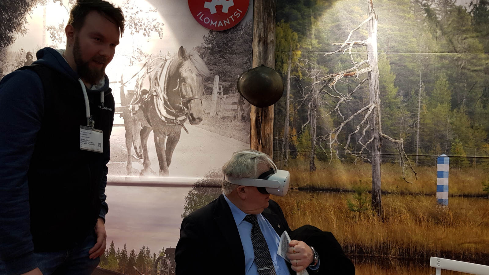 Kuvassa on taustalla valokeilassa Suomen hevonen vetämässä kärryjä. Toisessa osassa seinää on metsämaisemaa. Vasemmalla on Arto Väänänen neuvomassa tuolilla istuvaa Kyösti Kakkosta, joka katsoo virtuaalilaseilla Sotatien informaatiovideota.