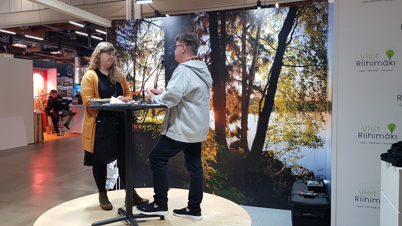 Kuvassa on vasemmalla Marja Hepoaho, jota haastattelee Miki Wallenius.  He seisovat korokkeen päällä.  Oikealla on seinaa, jossa lukee Riihimäki useassa kohtaa.