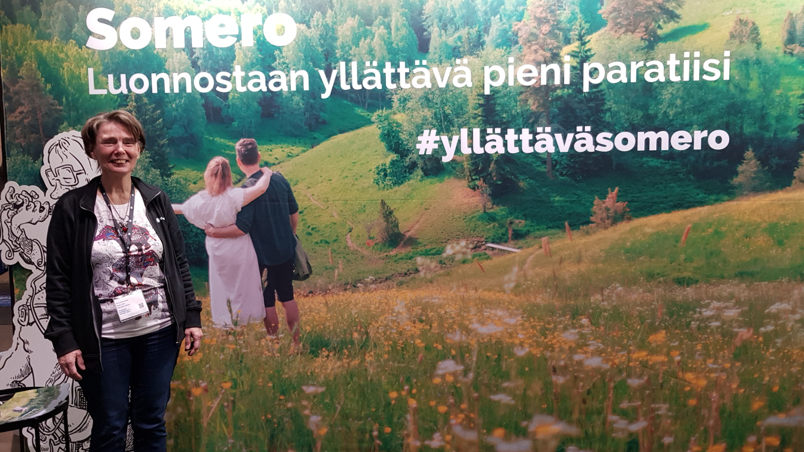 Kuvassa on kumpuilevaa kesämaisemaa ja vasemmalla on nuoripari katsomassa sitä. Ylä osassa lukee teksti Somero -luonnostaan yllättävä pieni paratiisi, hastag yllättäväsomero.  Vasemmalla on seisomassa Olla-Riitta Mäkelä.  