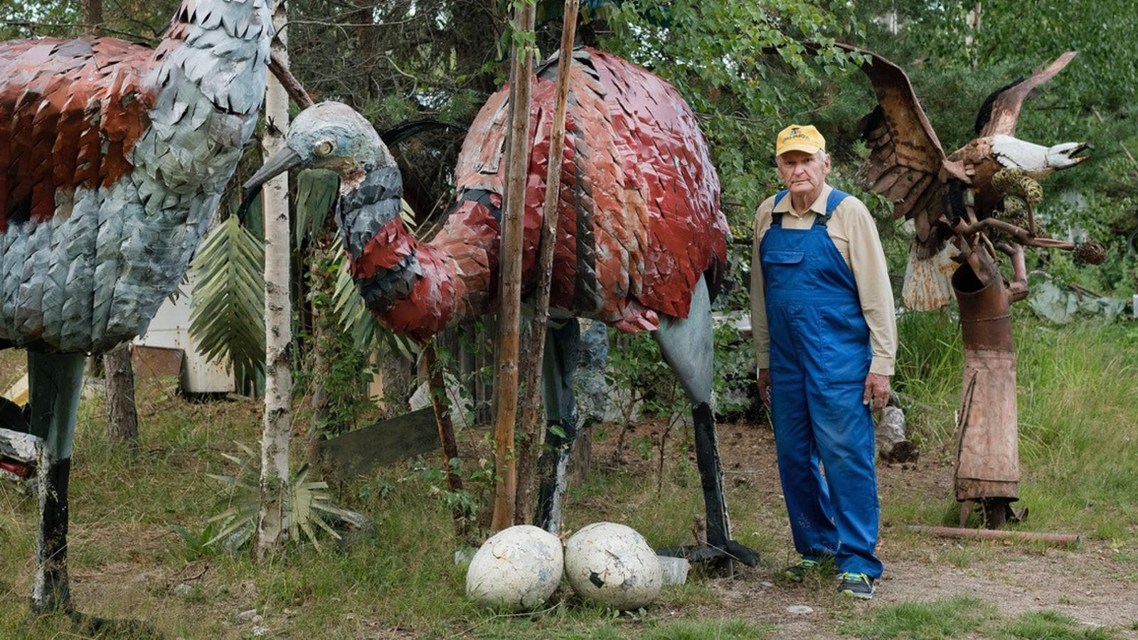 Kuvassa on Alpo Koivumäki sinisessä haalarissa seisomassa suurten metallista tehtyjen isojen strutsien ja kannon päässä olevan kotkan keskellä. Maassa on kaksi isoa vaaleaa strutsinmunaa.