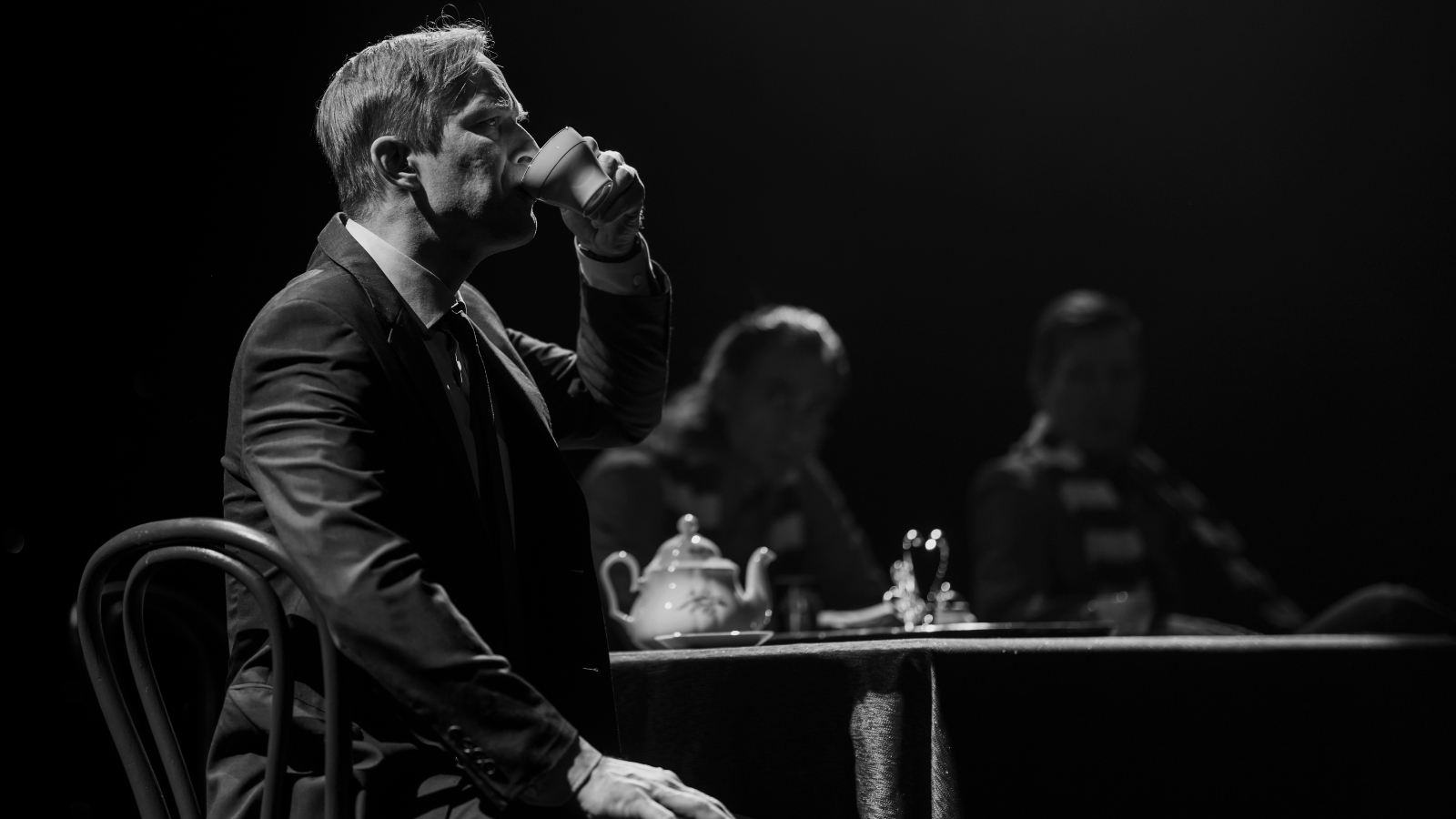 Kuvassa on Mikko Pörhölä juomassa teetä kahvilapöydän ääressä.  Kuva on musta-valkoinen.