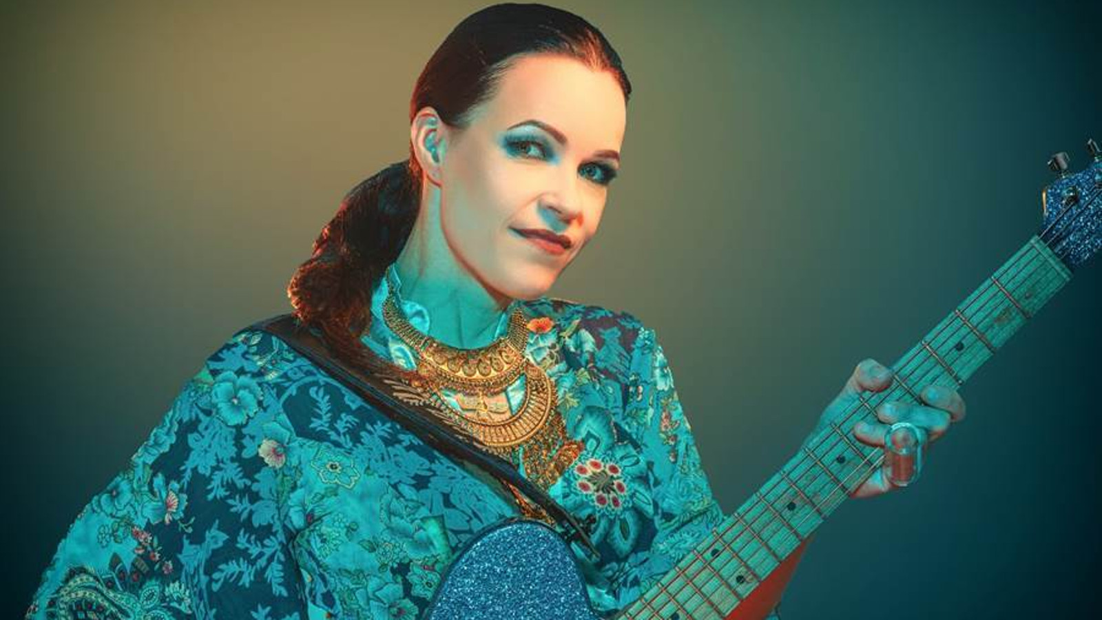 Kuvassa on Erja Lyytinen puolivartalokuvassa kitara kädessään.  Hänellä on sini-turkoosikuvioinen väljä pusero ja tausta on vihertävän sininen.