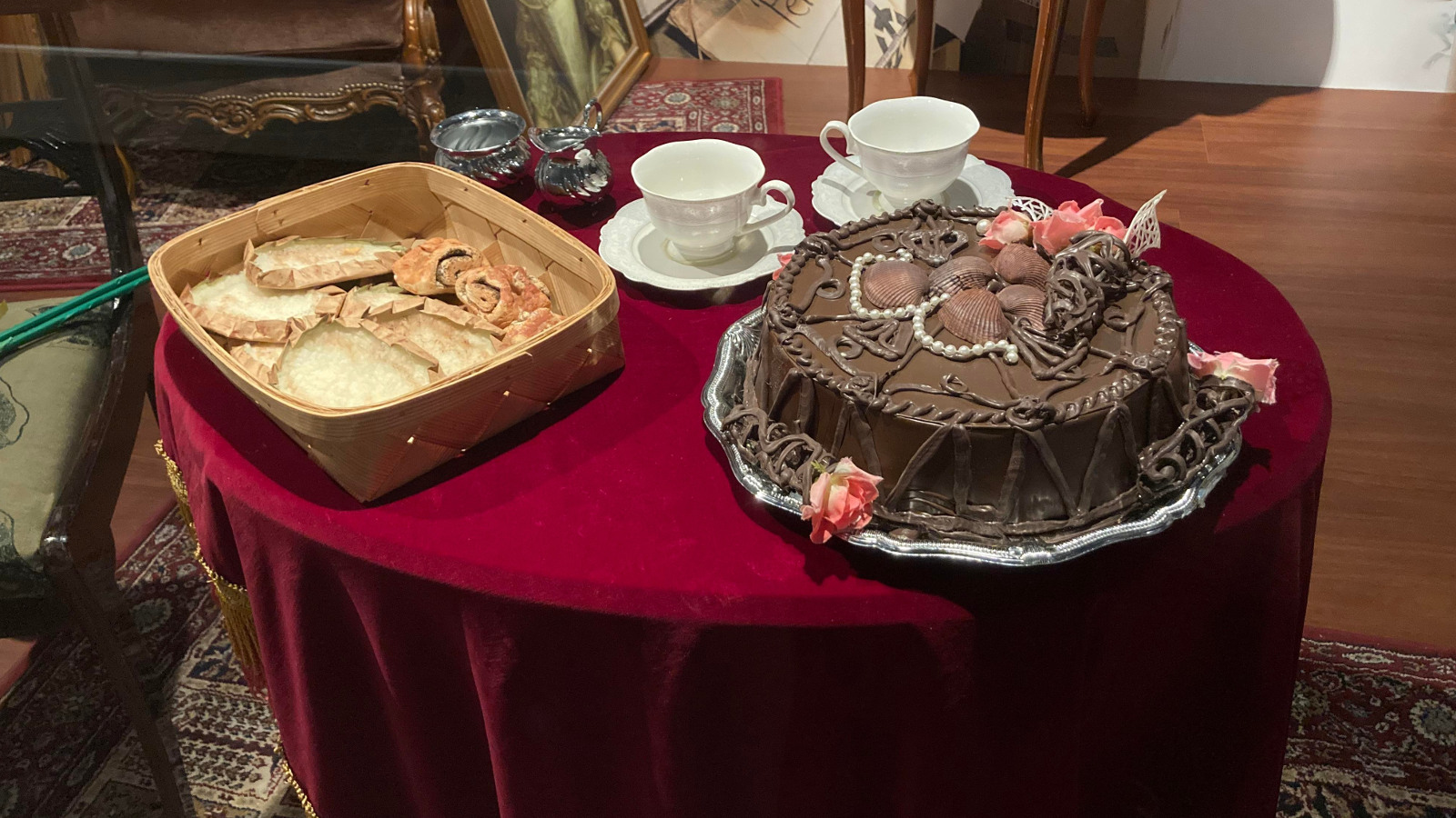 Kuvassa on punaisella samettisella pöytäliinalla oleva pyöreä pöytä, jolla on suklaapäällysteinen koristeltu täytekakku, piirakkakori ja kaksi kahvikuppia.