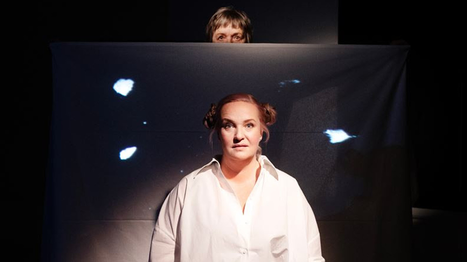 Kuvassa on tummalla taustalla eräänlaista kaksoiskuvaa, jossa on etualalla Nelly Juulia Kärkkäinen valkoisessa paitapuserossa. Takaa näkyy toinen kuva, josta näkyy puolet Gita Oksasen kasvoista.