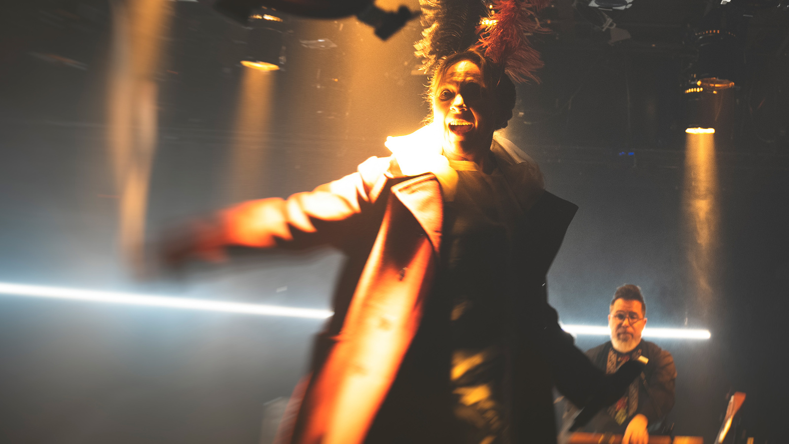 Kuvassa on Suvi-Sini Peltola ruskehtavassa takissa, johon osuu taustavaloa ja hän näkyy epätarkasti. Takana näkyy alhaalla kosketinsoittaja ja valopylväitä.