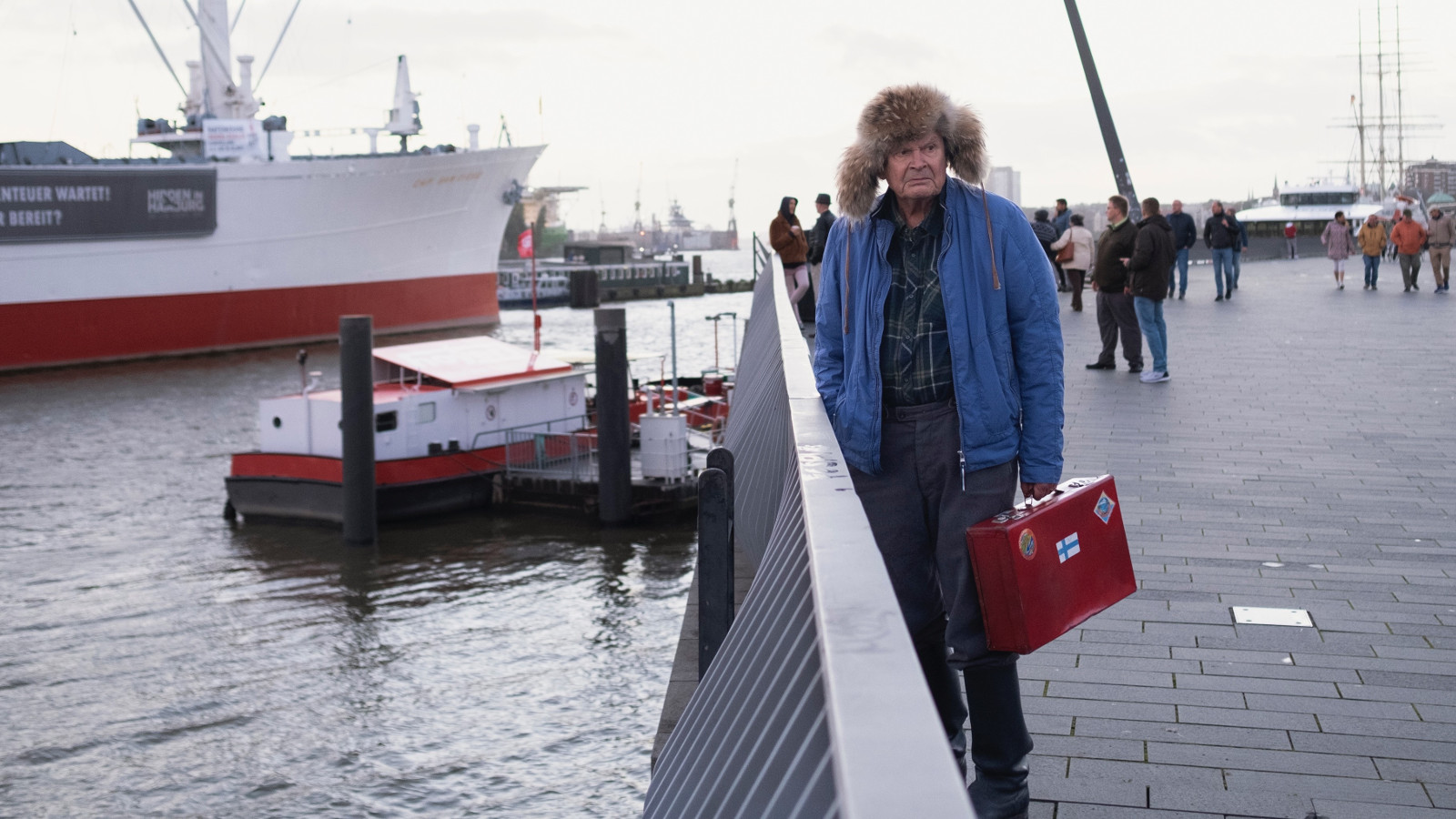 Kuvassa on oikealla Heikki Kinnunen sinisessä pusakassa ja karvahatussa satamassavesialtaan vieressä aidan luona.  Veden toisella puolella on puna-valkoinen laiva.