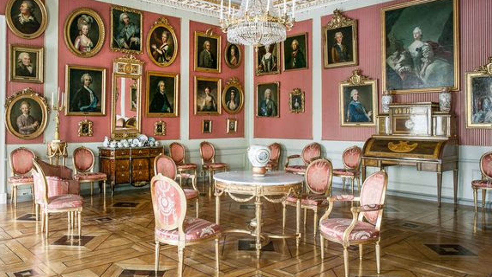 Kuvassa on etualalla kustavilaistyyliset valko-punaiset huonekalut ja vaaleanpunaisilla seinillä runsaasti pienikokoisia tauluja.  Katossa on on reunoilla koristeelliset ornamentit.