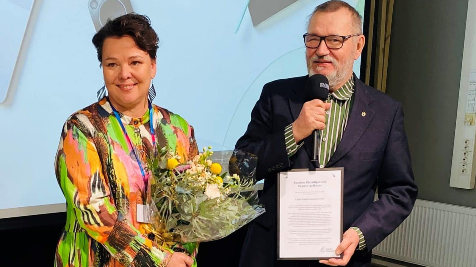 Kuvassa ovat Minna Kurttila värikkäässä mekossa ja kukat kädessään ja Erkki Kaila kunniakirja kädessään.  