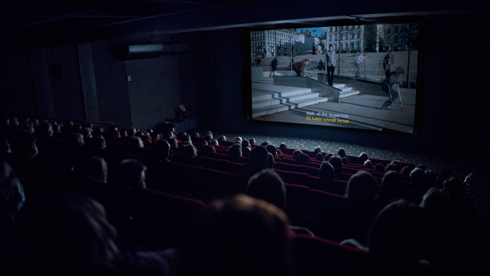 Kuvassa on pimeä elokuvasali, josta näkyy juuri ja juuri yleisöä.  Valkokankaalla näkyy näytettävä elokuva.