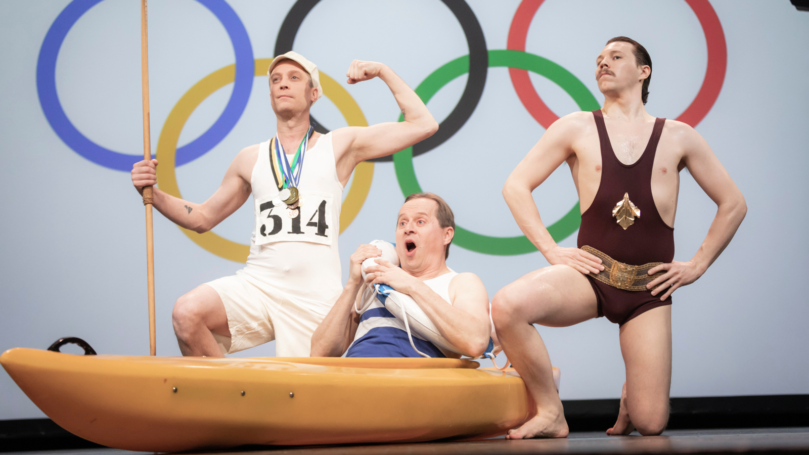 Kuvan tausta on valkoinen ja siinä on isot olympiarenkaat. Edessä on kolme miestä vanhanaikaisissa voimisteluvaatteissa ja he pullistelevat lihaksiaan.