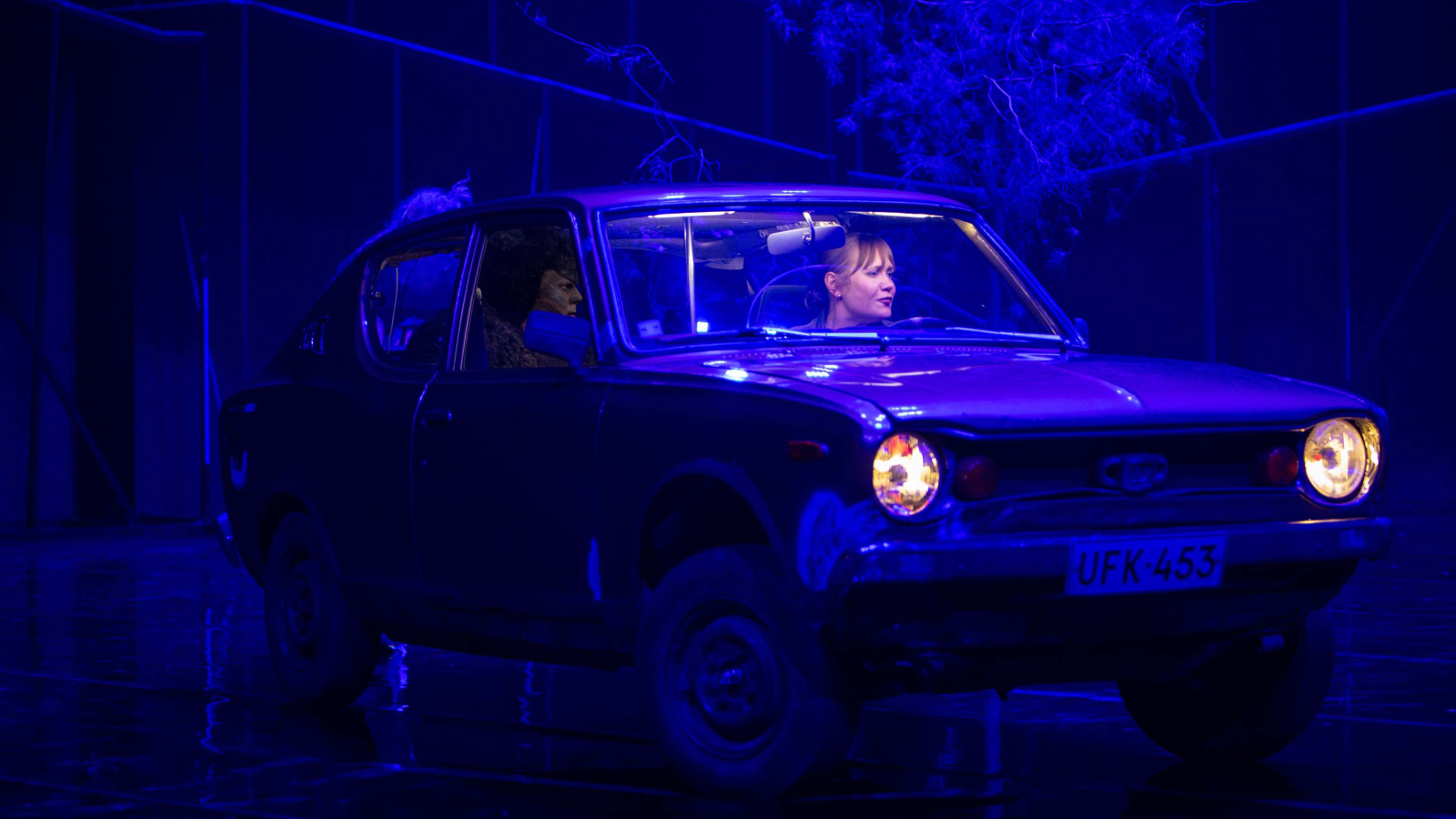 Kuvassa on Ella Lahdenmäki sinisen Toyota 100A:n ratissa. Kuva on tumman ja sinisen sävyinen.