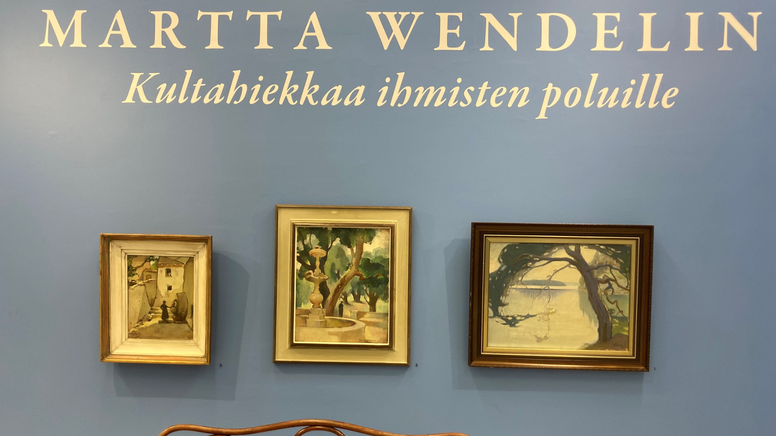 Kuvassa on kolme pientä taulua harmaalla seinällä ja näyttelyn nimi Martta Wendelin Kultahiekkaa ihmisten poluille.