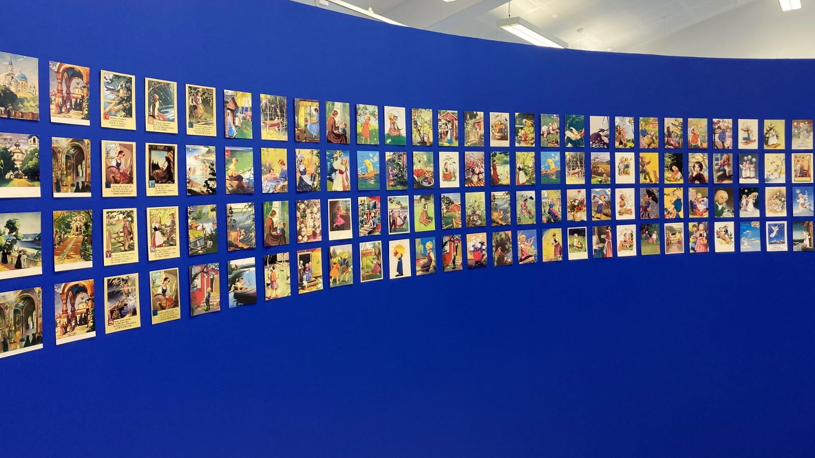 Kuvassa on sinisellä seinäll riveittäin runsaasti postikortteja.