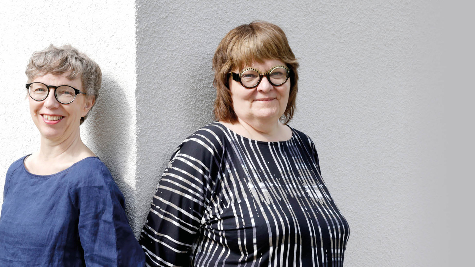 Kuvassa ovat Jaana Semeri sinisessä puserossa ja Leena Virtanen musta-valkoraidallisessa puserossa.  He nojaavat seinään ja välissä on vaalean seinän pieni kulma.