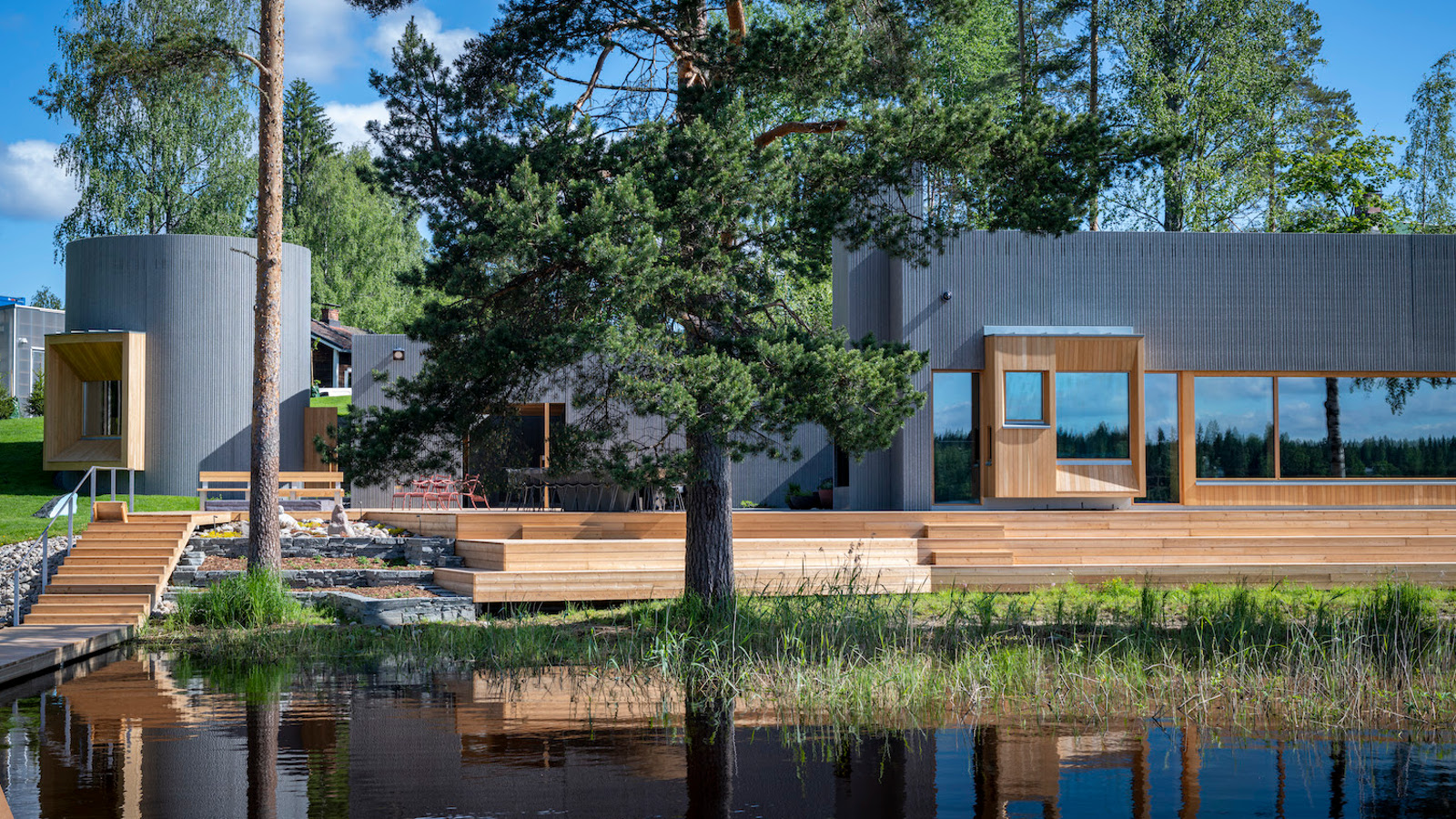 Kuvassa on Taidesaunan mustat rakennukset ja puun väriset laiturirakennelmat järven rannassa. Sauna on mäntyjen keskellä.