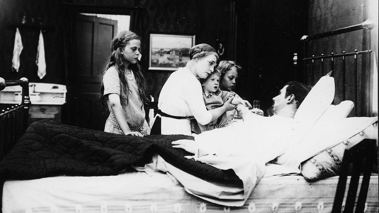 Kuvassa on mies makaamassa sairaana sängyssä ja sängyn vieressä ovat vaimo istumassa pienin lapsi sylissään. Takana seisoo tytär ja toinen kurkkii äidin takaa.