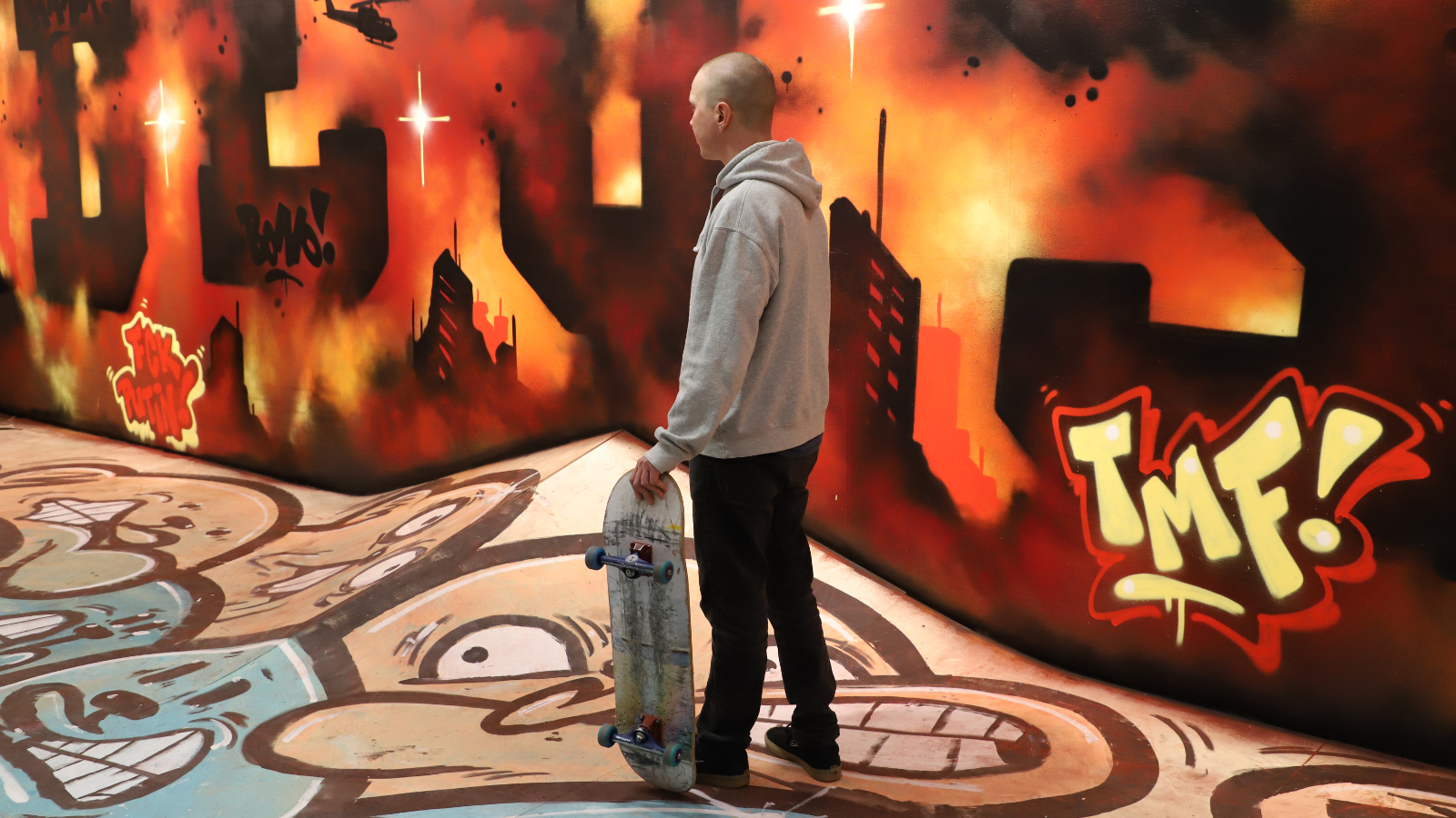 Kuvassa on aulatilassa värikkäitä graffitteja lattialla ja seinillä. Keskellä seisoo nuori mies skeittilauta kädessään. 
