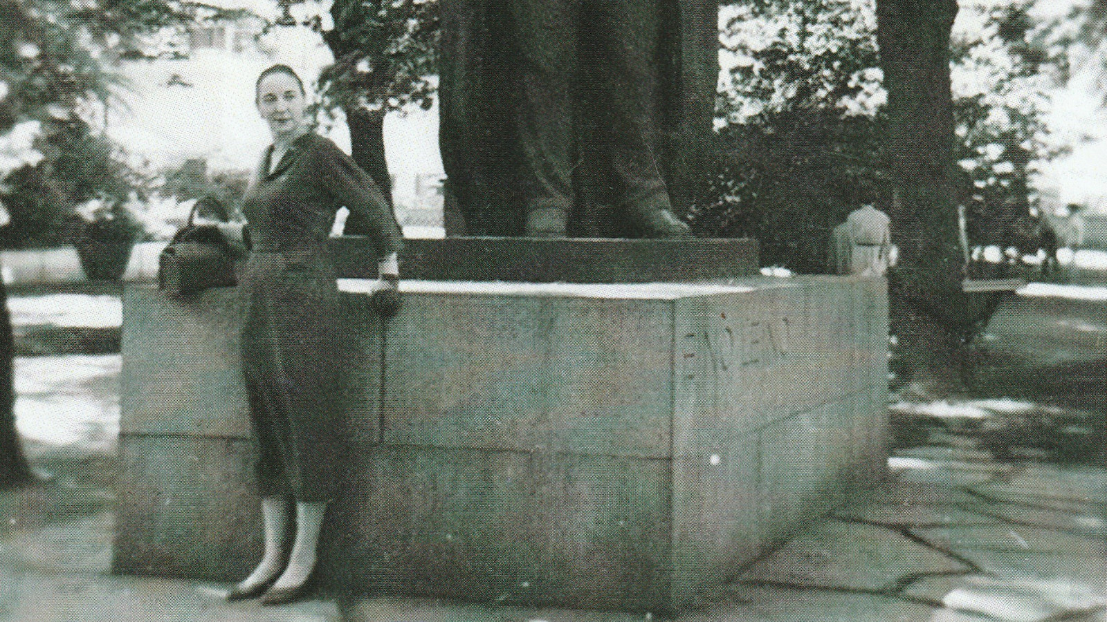 Kuvassa on Helena Eeva ison patsaan juurella seisomassa.  Hänllä on kapea musta puolihame päällään. Kuva on musta-valkoinen.