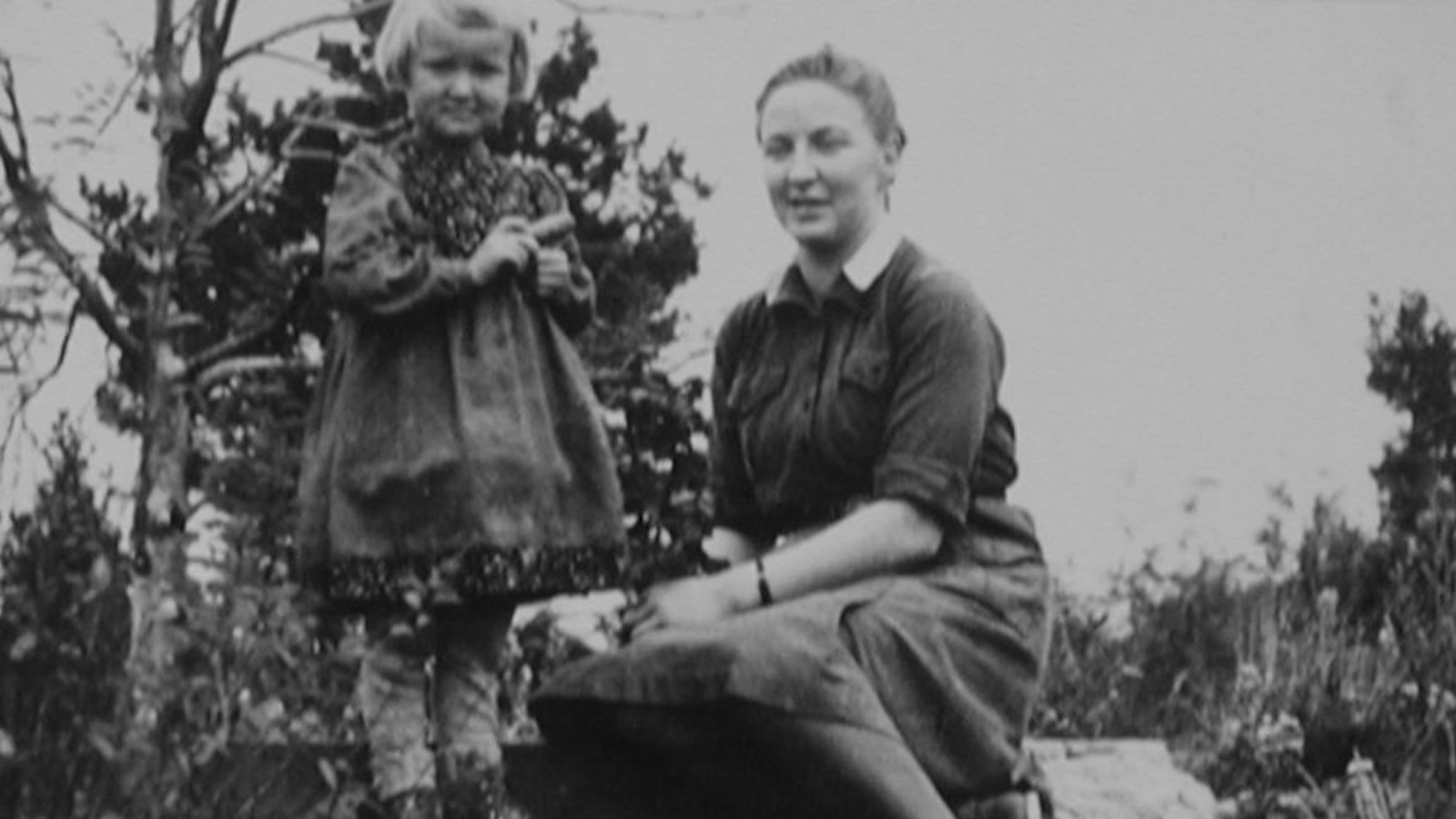 Kuvassa on Helena Eeva istumassa kahden betoniportaan päällä ja hänen vieressään on pikkutyttö mekossa. Kuva on musta-valkoinen.