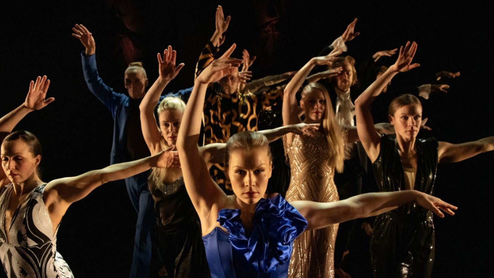 Kuvassa on joukko tanssijoita kädet ylhäällä ja edessä on tanssija sinisessä puvussa.