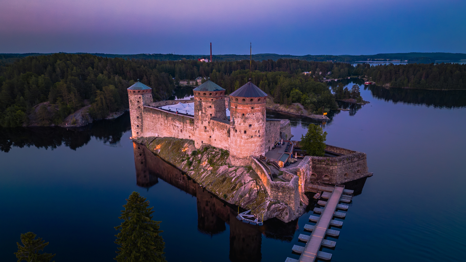 Kuvassa on Olavinlinna sinisävyinen ilmakuva iltahämäräisestä Savonlinnasta, jossa linna on veden keskellä ja taustalla näkyy metsää ja kaupunkia. Edessä on laituria, jossa on paikkoja veneille.