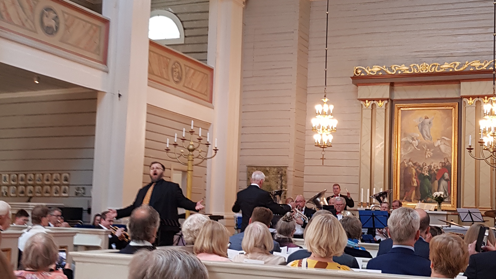 Kuvassa on Mika Nisula laulamassa kädet levitettyinä ja taustalla on Töölö Brassin soittajat.  Molemmin puolin näkyy yleisöä istumassa selin.