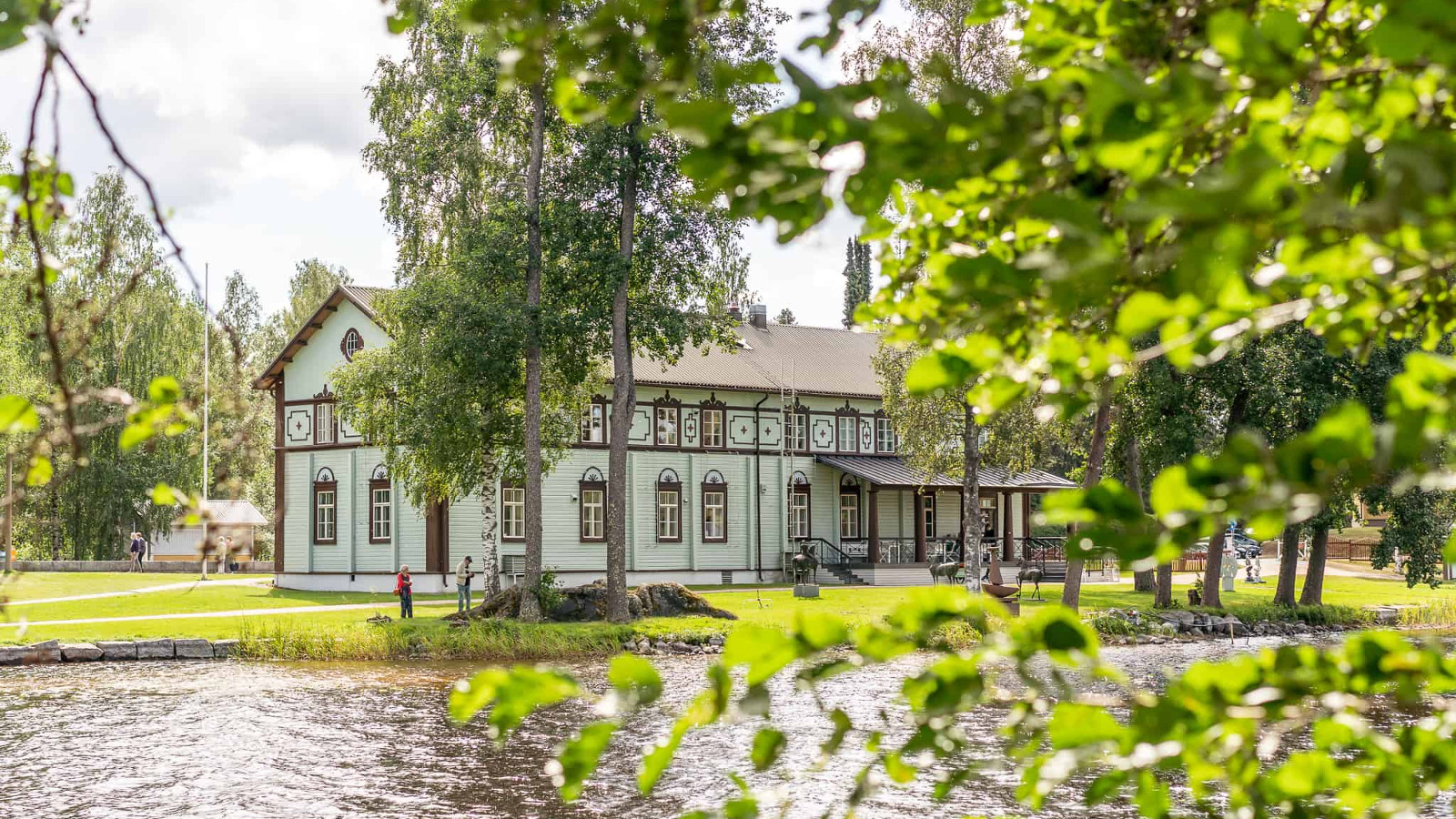 Kuvassa on Taidekeskus Salmelan päärakennus veden ääreltä kuvattuna.  Rakennus on turkoosi ja siinä on ruskeat ikkunapuitteet.