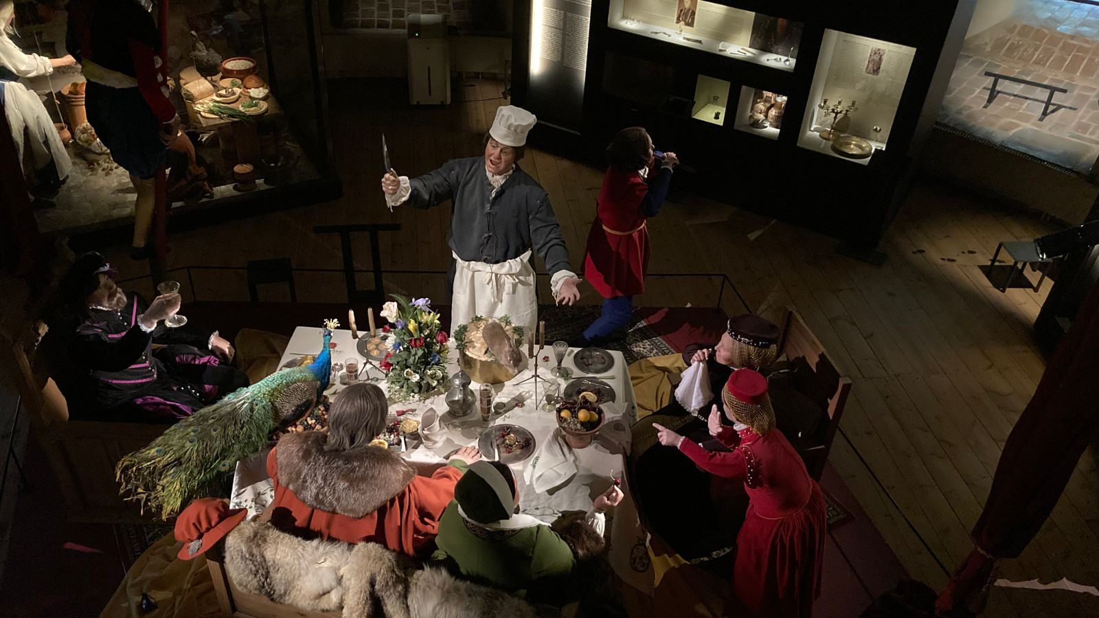 Kuvassa on katettu pöytä ja sen ympärillä ruokailijoita sekä kokki selostamassa ruuista. Henkilöt ja ruoka ovat nukkeja ja ruoka keinotekoista.