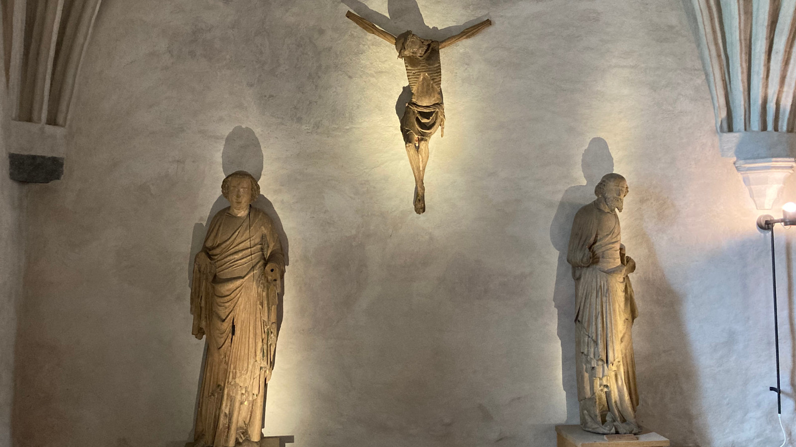 Kuvassa on harmaalla seinällä alempana kaksi pyhimyspatsasta ja ylempänä seinäveistos Kristus ristillä.