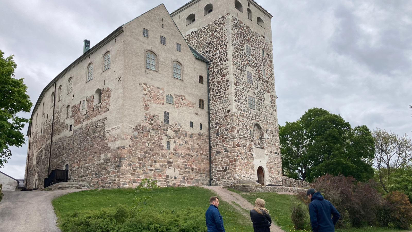 Kuvassa on harmaa Turun linna kuvattuna yläviistoon ja vihreää nurmikenttää.  Alhaalla näkyy kaksi miestä ja nainen katsomassa linnaa.