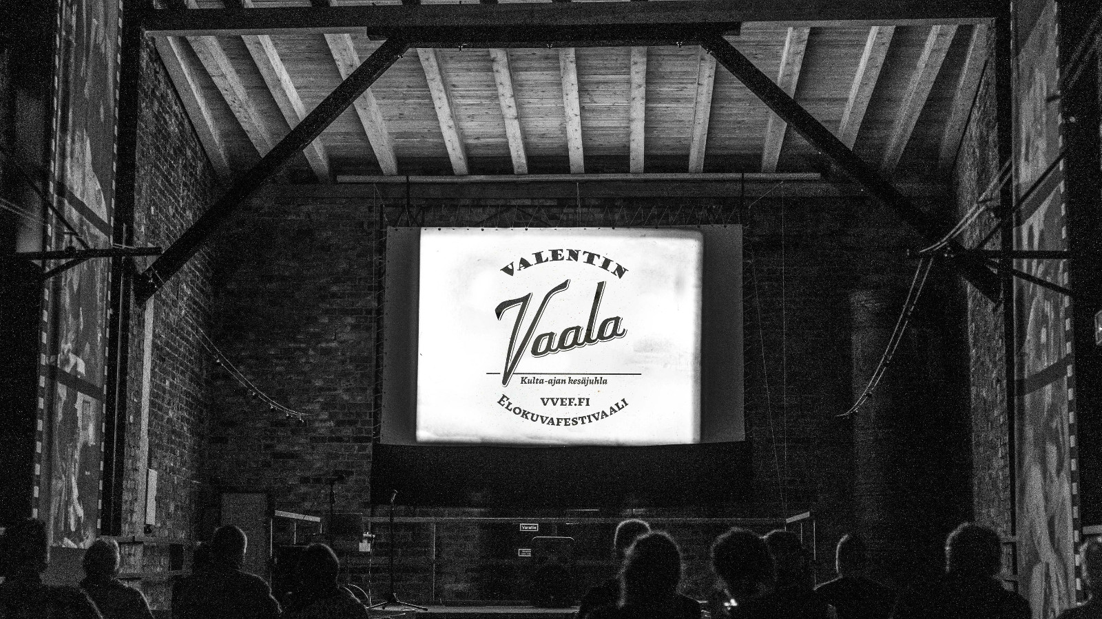 Kuvassa on vanhan ladon katsomo ja lautanäyttämö ja sen keskellä on pieni juliste, jossa lukee Valentin Vaala elokuvafestivaali. Lato on harmaa.