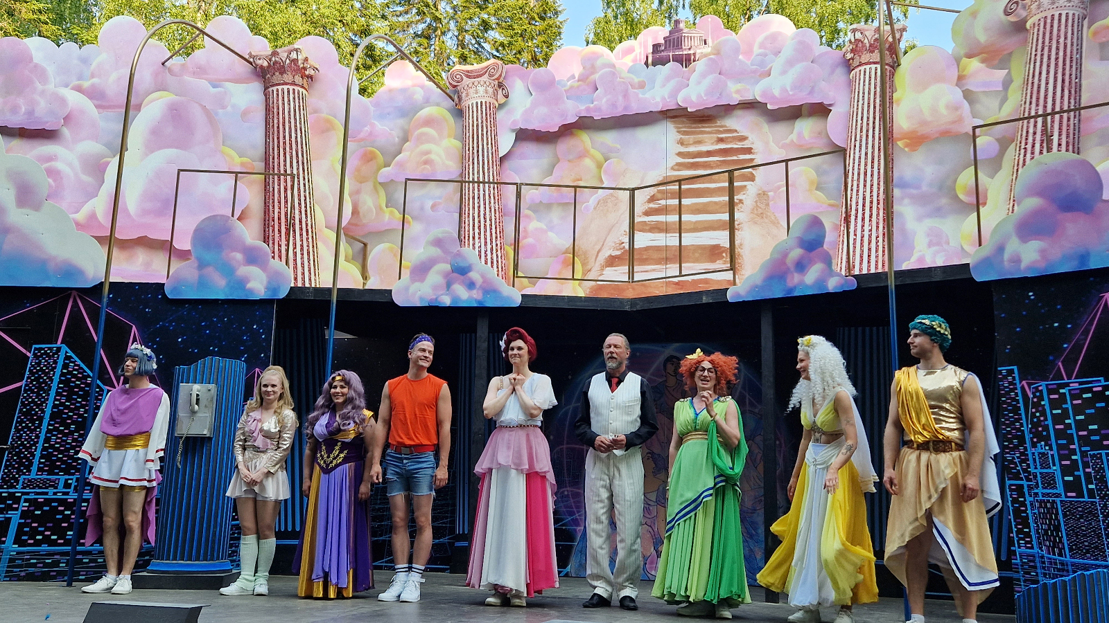 Kuvassa ovat Xanadun näyttelijät lavalla kiittämässä yleisöä. Taustalla on vaalean puna-sinisiä sokeritoppapilviä sekä antiikkisiä pylväitä.