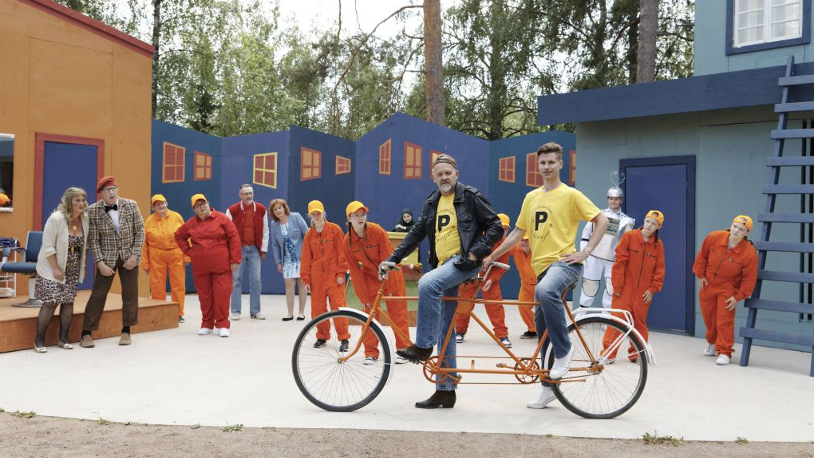 Kuvassa on etualalla kaksi miestä tandempyörällä ja taustalla ovat näyttelijät rivissä katsomassa heitä.