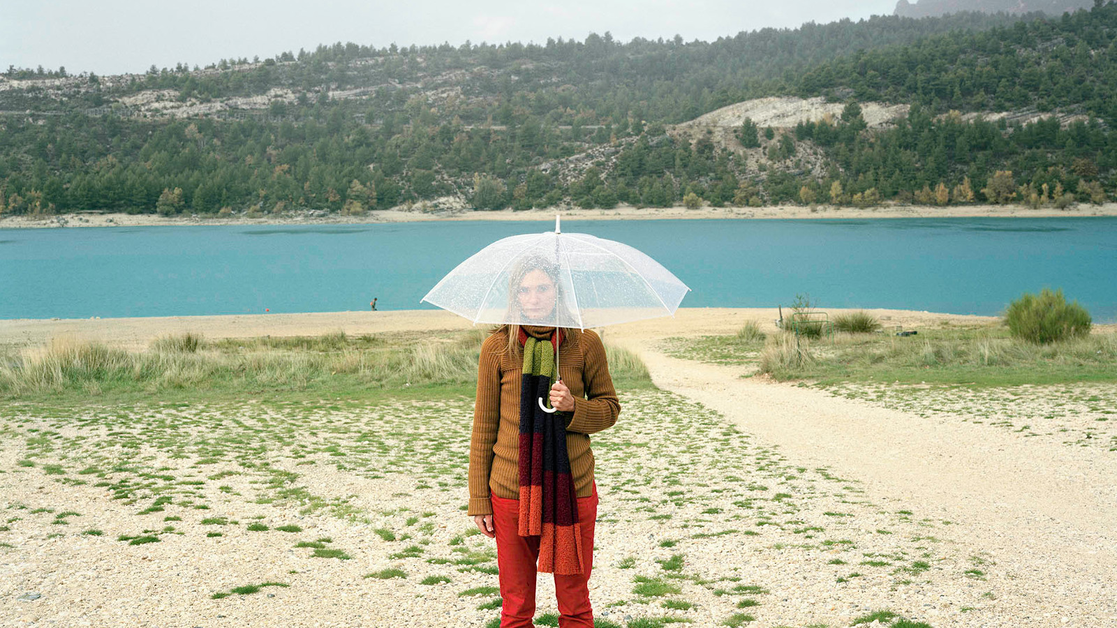 Kuvassa on nainen hiekkarannalla ja hänellä on sateenvarjo. Taustalla on sinistä vesistöä ja metsää.