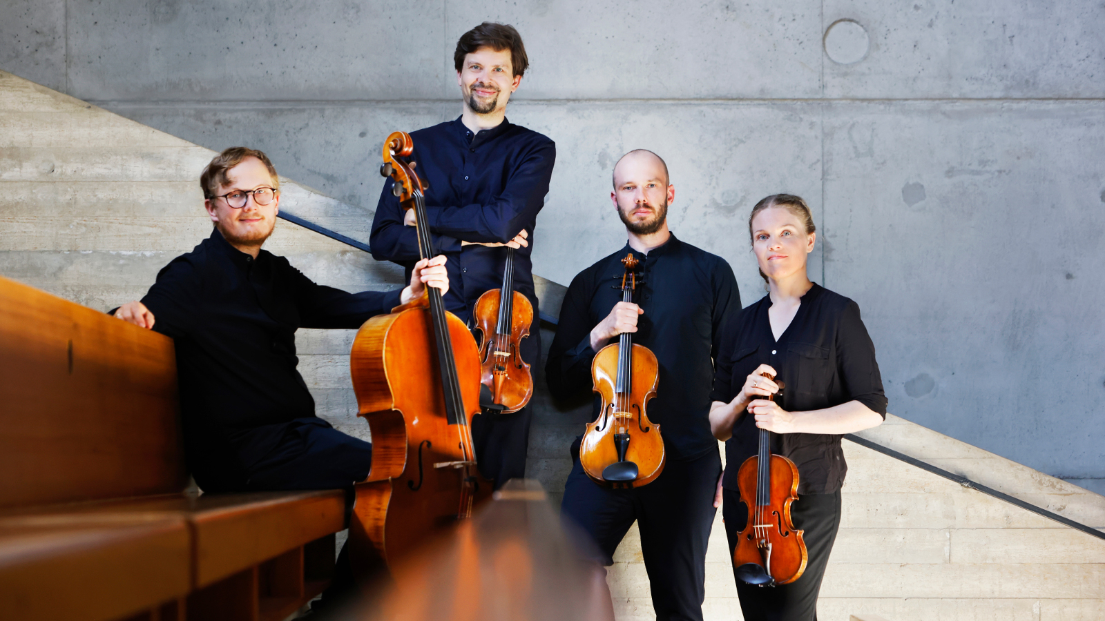 Kuvassa Kamus-kvartetin neljä soittajaa, joista vasemmalla oleva mies istuu ruskealla sohvalla ja pitää kädellään pystyssä selloa. Hänen vieressään oikealla seisoo mies viulun kanssa ja hänen vieressään mies myös viulun kanssa. Oikealla on seisoo nainen viulun kanssa.