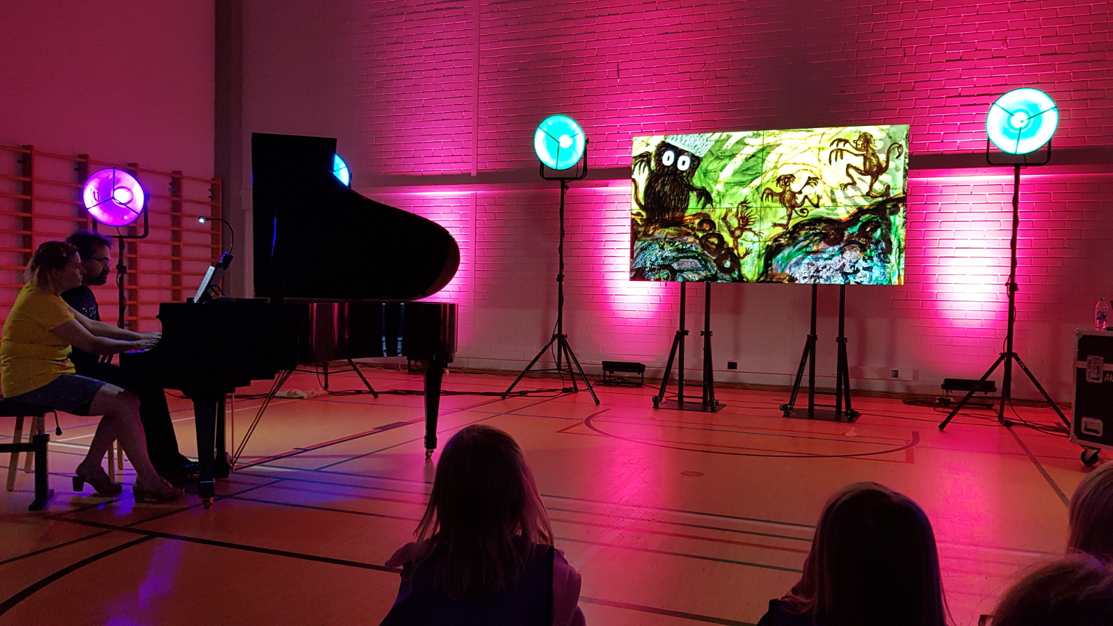 Kuvassa on tummassa salissa vasemmalla flyygeli ja sitä soittamassa Liisa Malmivaara ja Tuomas Turriago. Keskellä värivalojen keskellä on näyttö, jossa on punertavalla pohjalla iso kuningashahmo ja vieressä peikkoja.