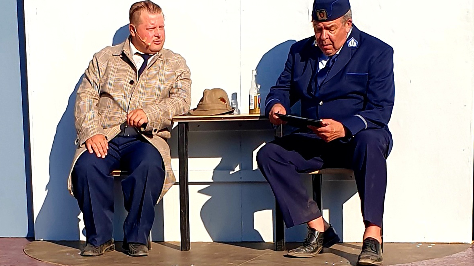 Kuvassa istuu vasemmalla Tenu vaalean ruskeassa popliinitakissa ja oikealla Reinikainen sinisessä poliisipuvussa. Välissä on pöytä.