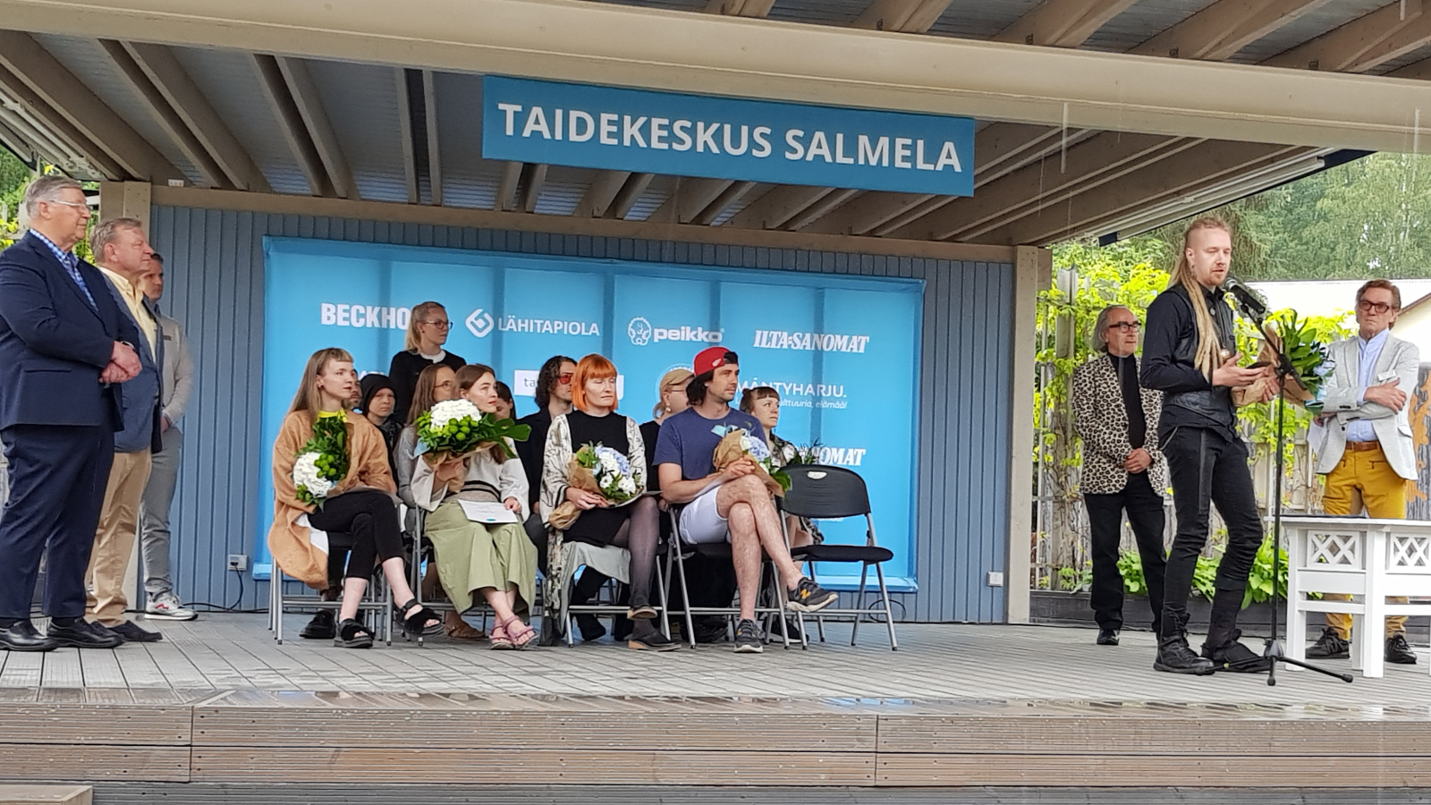 Kuvassa ovat keskellä näyttämöä istumassa nuorten taiteilijoiden näyttelyluokka ja vasemmalla tumman sinisessä puvussa Markku Häkkänen ja Markku Lankinen ja oikealla mikrofonin ääressä puhumassa Sakari Vinko mustissa vaatteissa.  Taustalla Esa Saarinen leopardikuvioisessa takissa ja Tuomas Hoikkala vaaleassa takissa ja keltaisissa housuissa.