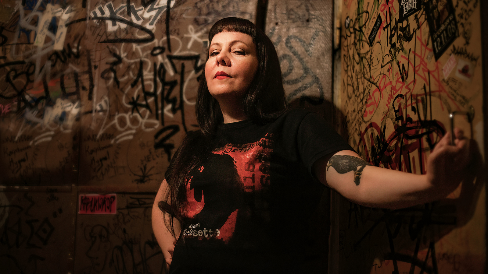 Kuvassa on Tiina Käpylä ja hänellä on tummat, pitkät hiukset sekä musta-punainen puku. Taustalla on graffitiseinää.