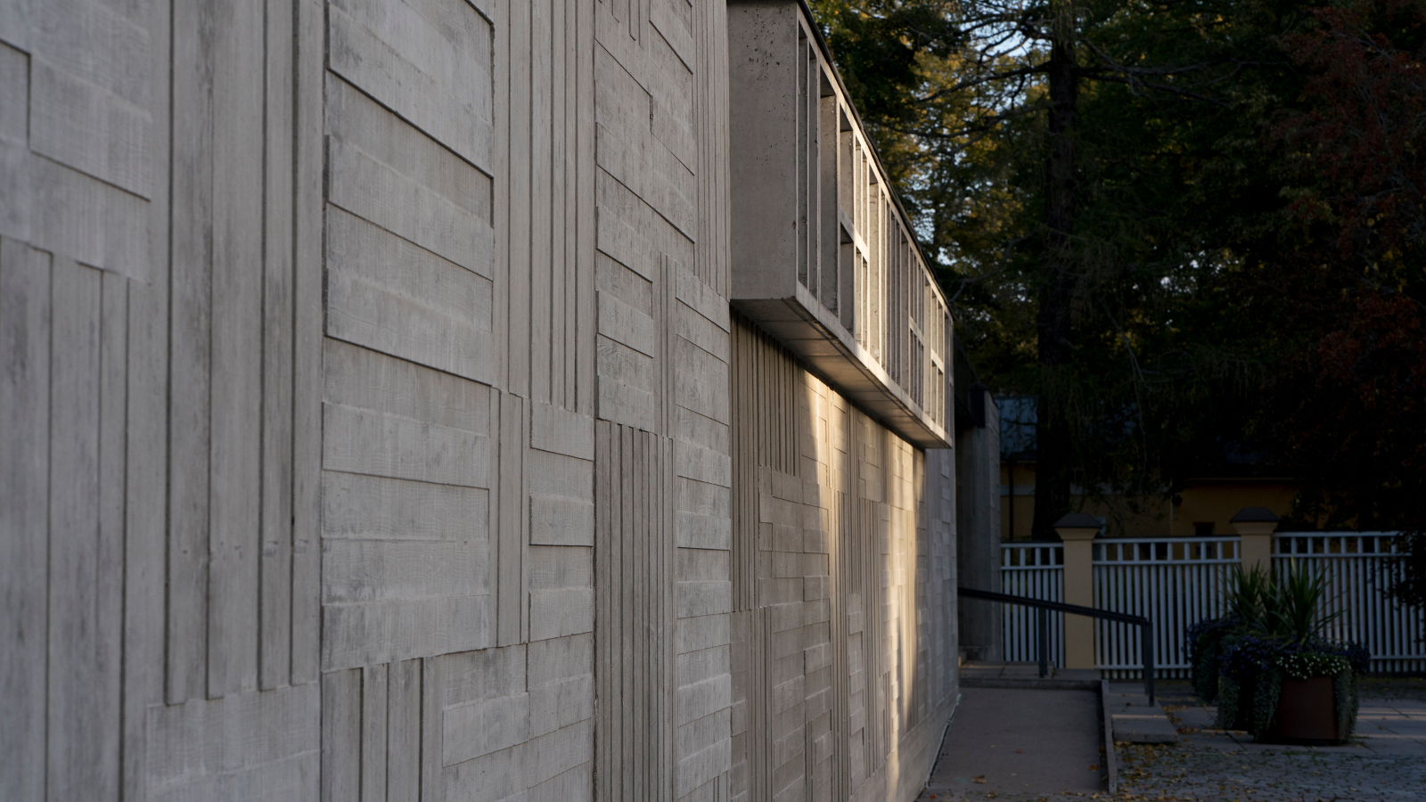 Kuvassa on harmaata betonista ulkoseinää, jossa pysty- ja vaakasuoria pintakohotuksia.  Oikealla ylhäällä on pieni hylly.
