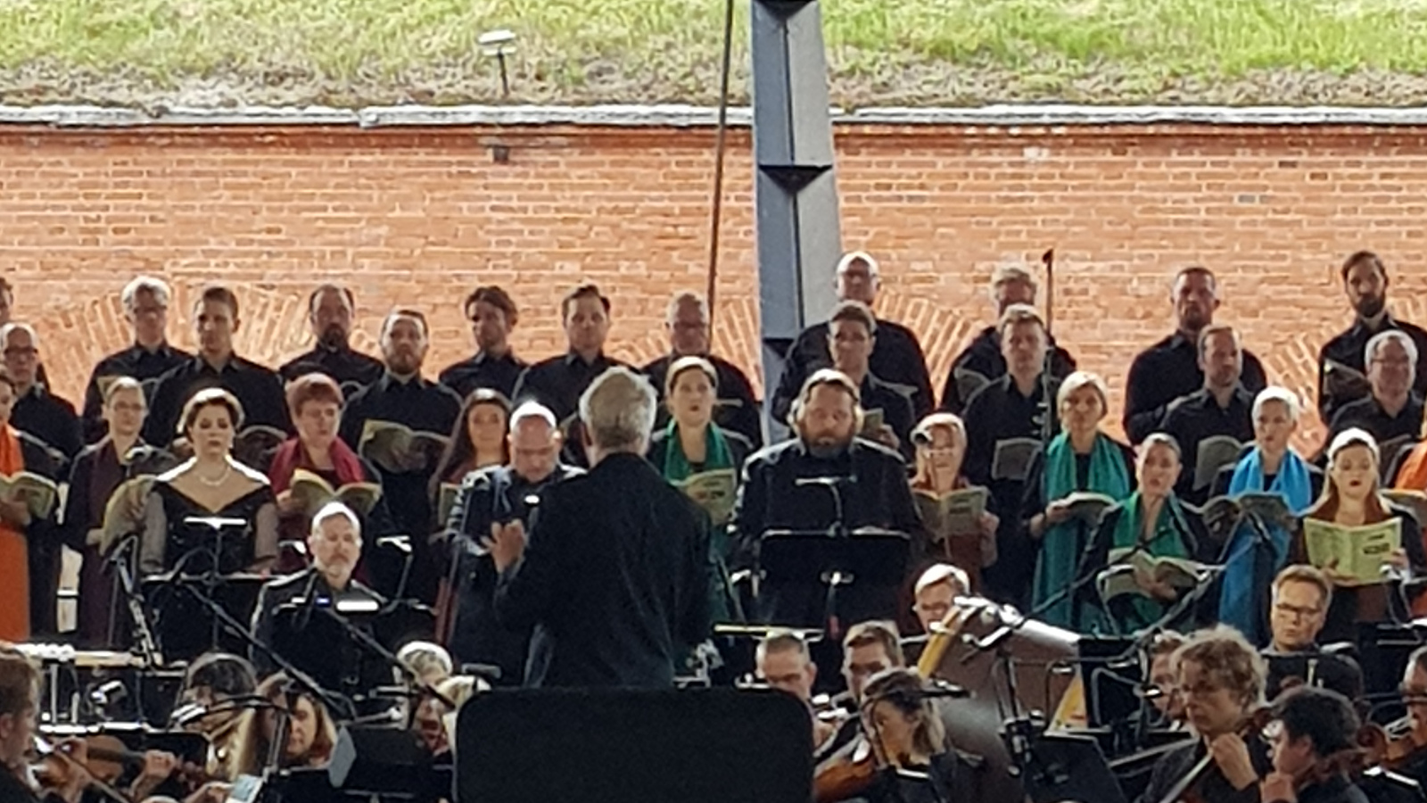 Kuvassa on Hannu Lintu selin johtamassa orkesteria ja hänen sivuillaan on soittajia. Edessä on vasemmalla solistit vasemmalla sopraano Silja Aalto, tenori Mika Pohjonen ja basso Matti Turunen. Taustalla on kuorolaulajia sekä tiilimuuria.