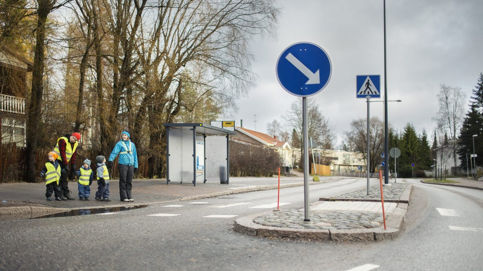 Kuvassa on vasemmalla lähdössä ylittämään katua keltaisessa liivissä nainen, jolla on kaksi pientä keltaliivistä lasta kädessään. Vieressä nainen vaalean sinisessä heidastinnauhaisessa takissa ja pitää kädestä pientä lasta. Keskellä kuvaa on liiktenteen jakaja.