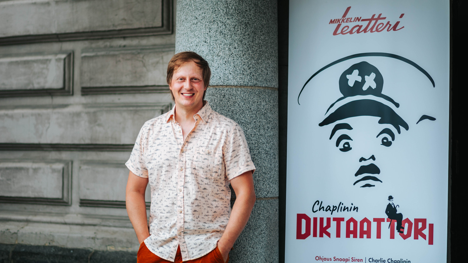 Kuvassa on Juha Pulli, joka nojaa isotiiliseen Mikkelin teatterin seinään.  Oikealla puolella on Diktaattorin juliste, joka on musta-valkoinen ja siinä on hitlermäinen graafinen kasvokuva sekä näytelmän nimi.