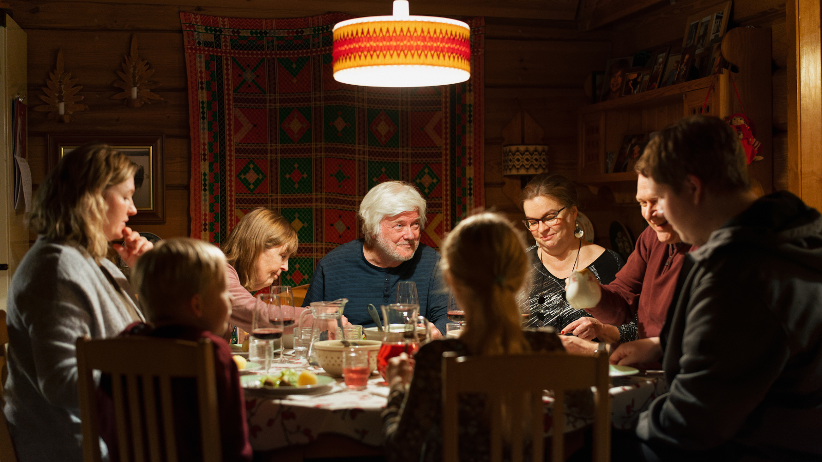 Kuvassa on keskellä pöydän ääressä vaalea hiuksinen vanhempi mies ja ympärillä eri-ikäisiä henkilöitä kuusi ja yksi lapsi syömässä.  Taustalla oleva seinä on ruskea.