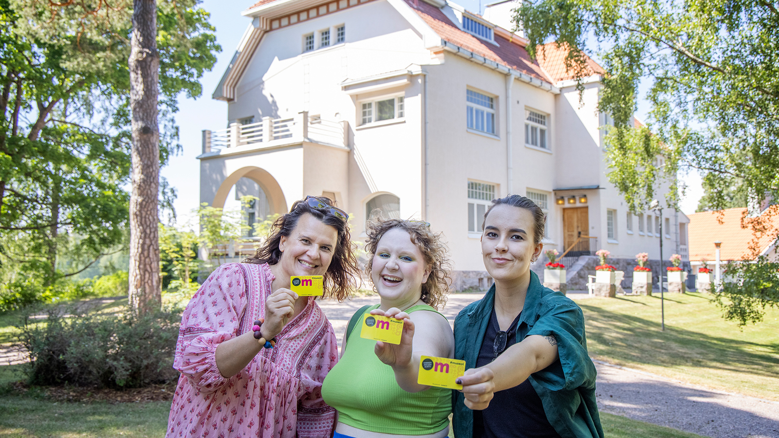 Kuvassa on taustalla Tamminiemen päärakennus, joka on vaalea jugend-talo.  Edustalla on kolme naista vierekkäin ja he näyttävät kaikki museokorttia.